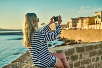 Fototapeta na wymiar Woman traveler tourist using smartphone, taking photo of sea view at sunset in summer day. Enjoying European, Famous popular touristic place in world. 