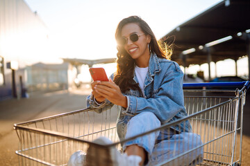 Young woman with phone sitting shopping cart having fun near color wall.  Blogging. Shopaholic, customer, sale concept.