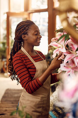 Happy black female florist arranging fresh flowers while working at flower shop. - obrazy, fototapety, plakaty