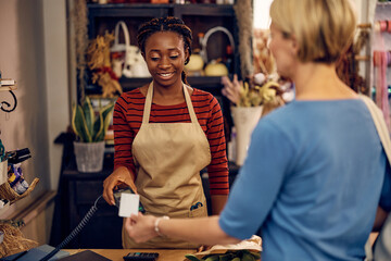 Happy African American florist receives credit card payment from her customer at flower shop.