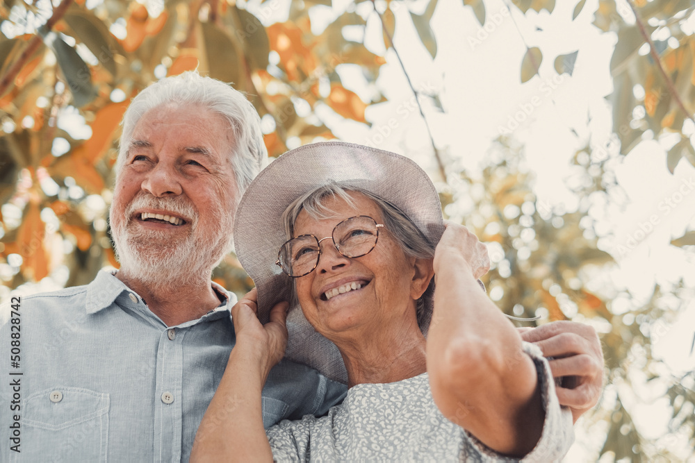 Sticker Elderly couple embracing in spring park outdoors having fun and enjoying together looking at the trees. Two old and mature people in love caring each other..