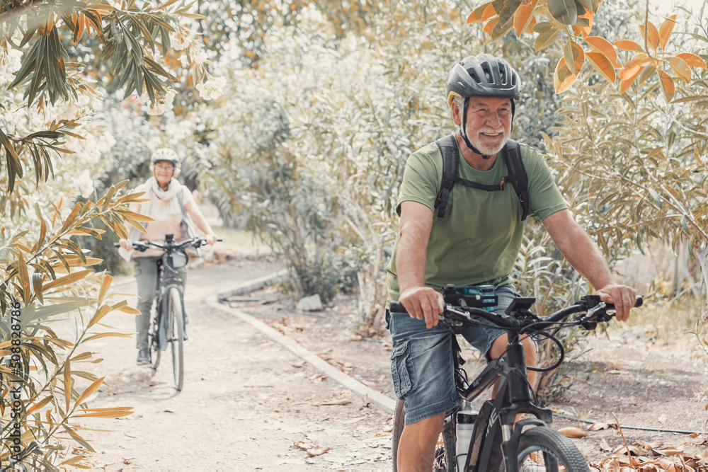 Sticker Two happy old mature people enjoying and riding bikes together to be fit and healthy outdoors. Active seniors having fun training in nature.