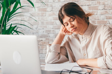 Young woman student sitting at desk full with books textbooks looking at computer screen to unfinished homework feels tired bored and unmotivated, girl loaded with tasks, work backlog overflow concept