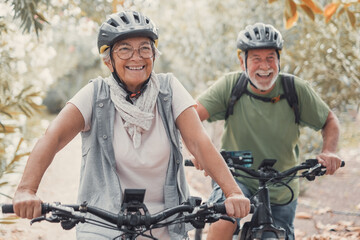Two happy old mature people enjoying and riding bikes together to be fit and healthy outdoors. Active seniors having fun training in nature. - obrazy, fototapety, plakaty