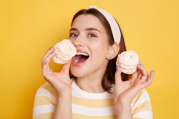 Indoor shot of attractive excited woman wearing t shirt and hair band standing with marshmallow in hands, feels hungry, wants to bite, posing with open mouth isolated on yellow background.