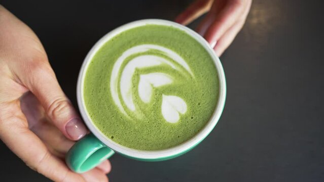 Female hands put cup of green matcha with latte art on black table. Barista serves matcha latte in cafe. Trendy matcha tea in green cup closeup