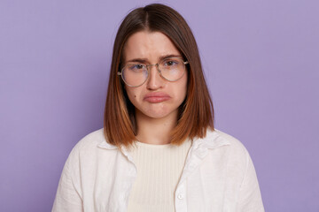 Obraz premium Portrait of beautiful dark haired young Caucasian woman wearing white shirt looking at camera with pout lips, being sad and upset, expressing sorrow, posing isolated over purple background.