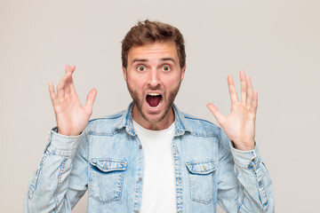 Young hipster male with stubble in a white t-shirt and light blue denim shirt. Emotion - a frightened scream, raised hands, an angry grin. On a light gray background