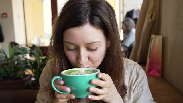 Young woman in cafe takes sip of matcha latte from green cup. Woman tastes trendy matcha tea and pleasantly smiles looking at camera