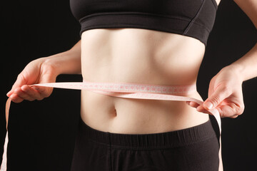 A woman measures the circumference of her abdomen on a black background. The concept of weight loss
