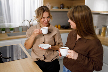 Happy middle aged woman enjoying coffee with her adult daughter at the kitchen
