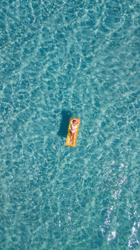 Aerial vertical view of woman relaxing and enjoying summer holiday vacation laying on a colorful inflatable lilo mattress on blue water surface. People enjoying ocean travel destination. Vertical