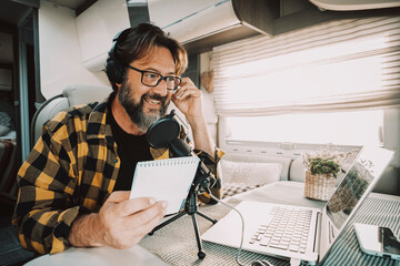 Modern adult man wokring with laptop and microphone inside a camper van in digital nomad job...