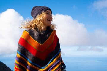 Colorful poncho clothes on happy pretty adult woman enjoying outdoor leisure activity against a blue sky background. Happy and cheerful people female portrait with winter hat - obrazy, fototapety, plakaty