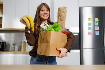 Cheerful young woman at the kitchen holding grocery shopping bag with vegetables and other product