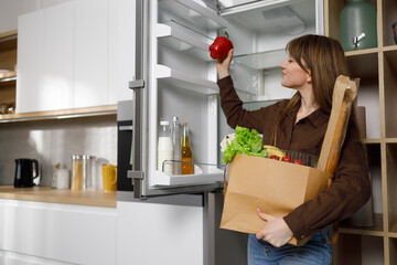 Woman with a paper shopping bag full of vegetables in the kitchen puts groceries in the fridge