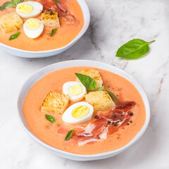 Spanish salmorejo soup, tradition Andalusian cold tomato soup topped with serrano ham (jamon), boiled egg, bread and pepper on the white background, square crop. Summer cold recipes