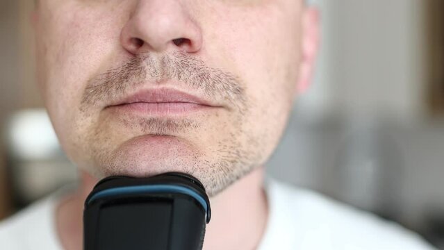 Handsome young unshaven man shaving with modern electric razor trimer while looking at camera, indoors. Daily routine male self-care. man shaves with an electric razor in the kitchen.