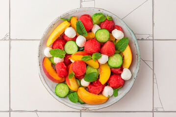 Summer fruit salad with mozzarella cheese, cucumber and mint in a plate on white tile background. top view