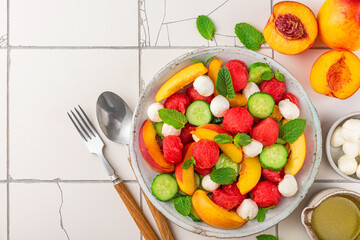 Summer fruit salad with mozzarella cheese, cucumber and mint in a plate with cutlery on white tile table. top view