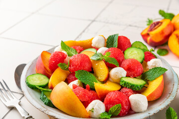 Fototapeta na wymiar Fruit summer salad made of fresh fruits, mozzarella cheese, cucumbers and mint on white tile table. Healthy diet food