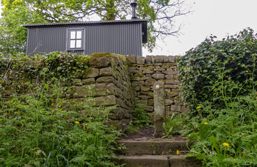 Iconic shepherds hut, sited behind an old local dry stone wall. Ancient  worn steps up, flanked with vegetation. Background of trees and sky. Landscape image with space for copy. Derbyshire. England. - 508852745