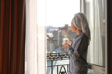 smiling young woman in nightwear and towel on head is holding cup of hot coffee or tea on balcony by the window in the morning. Concept of domestic lifestyle.