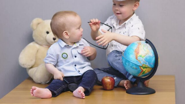 two little boys playing. elder brother putting glasses on toddler baby child. funny siblings having fun, educational concept, toy globe, teddy bear and apple. happy children games, school education