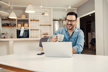 A man holding mug with beverage and smiling at the laptop form home.