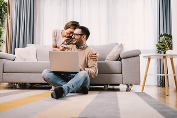 A happy couple hugs and uses a credit card for online shopping on a laptop at home.
