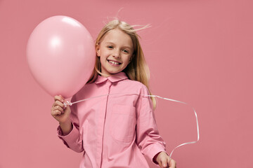 a happy, joyful school-age girl stands on a pink background with a big balloon in her hand and smiles broadly while looking at the camera.