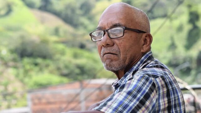 close-up of an elderly latin man with brown skin and glasses, standing with his hands on the railing of a balcony at home, observing nature and thinking about his long life. concept of retirement