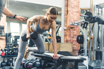 Fototapeta na wymiar Strong fit sporty caucasian woman in her 20s lifting weights with one hand and leaning on a bench with the other. Gym facility concept. High quality photo