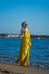 Arab woman with hijab and yellow Moroccan dress on the beach, enjoying her summer vacation. Walking along the shore. Vertical photo