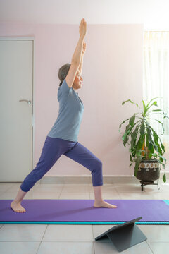 Full length shot of energetic mature woman making exercising and practicing yoga on mat while looking tablet for online lessons at home because of social distancing, wearing sports clothes.