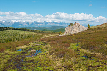 Obraz premium Atmospheric landscape with big rock on hillside with view to sunlit mountain vastness and high snowy mountain range under low clouds line on horizon. Impressive scenery with vast mountains in sunlight