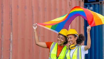 Young female African sisters in safety uniform and helmet show LGBT flag to support freedom and rights for gender equality next to red cargo or shipping container