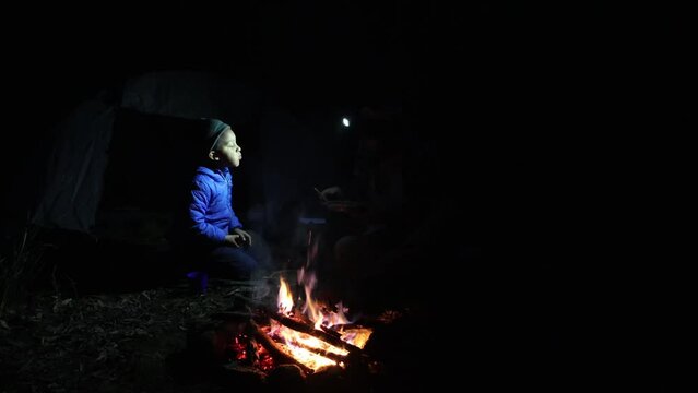 A man and his son sit around a campfire in the dark night in a forest with their torches shining on their food.