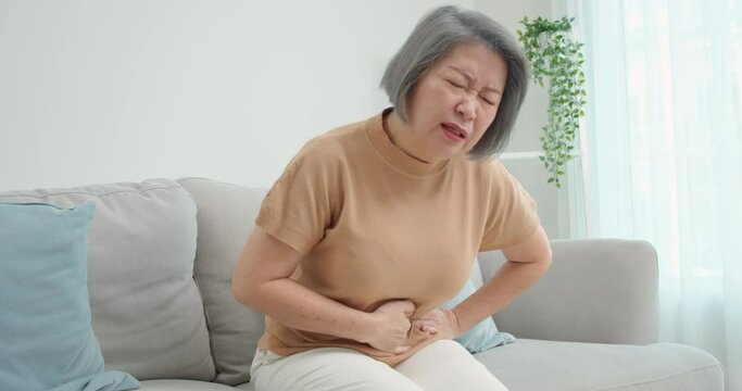 Beautiful elderly Asian woman sits on the sofa, hand clutching her stomach due to severe abdominal pain,diarrhea, gastritis, enteritis in the living room alone.People's health problems concept.