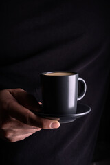 A man's hand holds a cup of hot espresso coffee against the background of a black T-shirt.