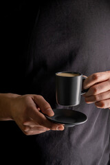 A man's hand holds a cup of hot espresso coffee against the background of a black T-shirt.
