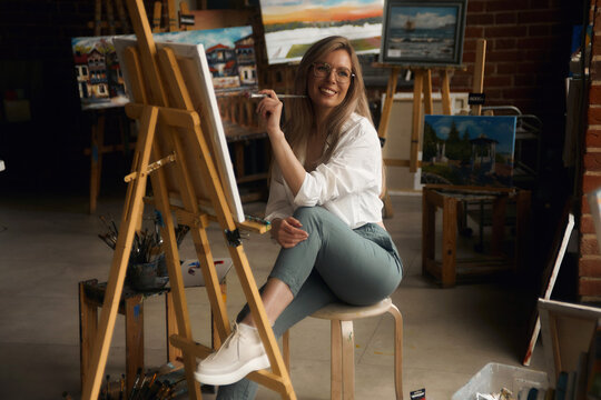 Inspired blond teenage girl with brush and palette smiling in studio interior. Creative schoolgirl enjoying her new hobby, still life painting, side view. Art concept