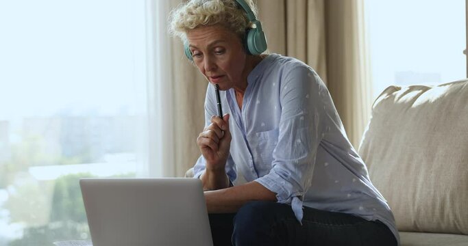 Middle-aged woman in wireless headphones sit on sofa use laptop listens audio course, gain new knowledge engaged in self-education. Older gen use modern tech for e-learning, refresher training concept