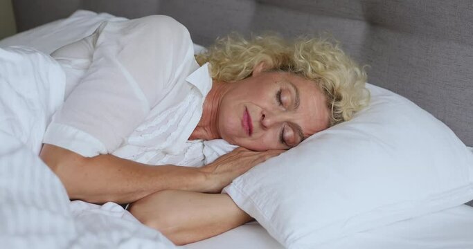 Close up beautiful older woman put head on soft hypoallergenic memory foam pillow sleeping lying alone in bed looks serene deep in dreams. Comfort orthopaedic mattress for quality healthy rest concept