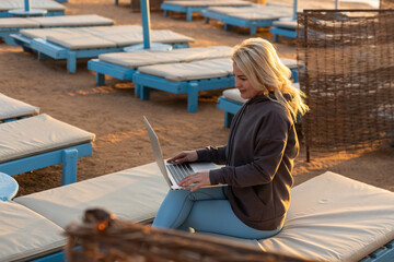 Close-up image of woman sitting on sandy beach and working on laptop, coding or answering e-mails.