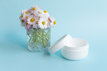 A bouquet of fresh flowers, daisies on green stems are in a glass with clean water and a white jar of cream on a blue background