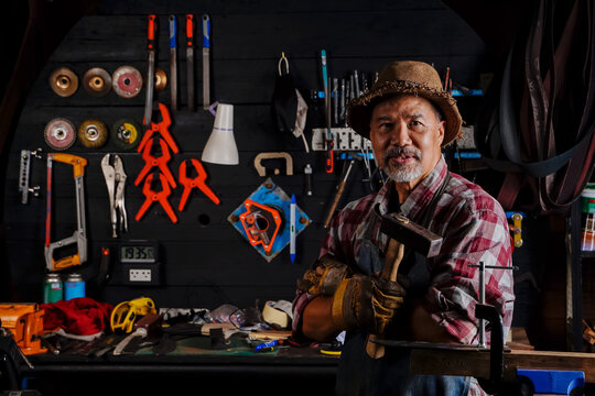 An elderly man with a blacksmith's beard stood proudly crossed his arms and held a hammer in the smithy.