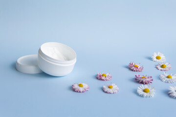 Fresh flowers, daisies are laid out in a corner, with a glass of clean water and a white jar with a cream cosmetic on a blue background and a free space for advertising.