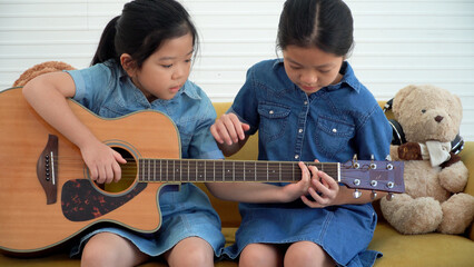 Asian teenage girl teach younger playing acoustic guitar sitting together on yellow sofa at home....