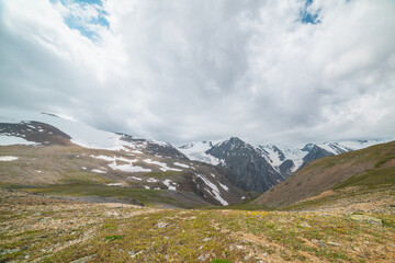 Fototapeta premium Scenic panoramic view from sunlit green grassy hill to high snowy mountain range with sharp tops and glaciers under gray cloudy sky. Colorful landscape with large snow mountains at changeable weather.